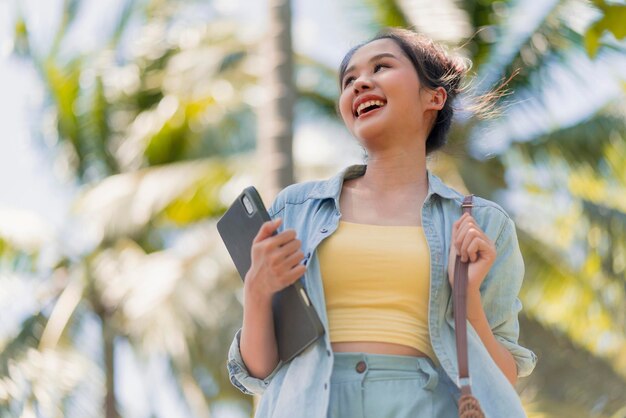 편안하게 웃고 있는 아시아 여성 디지털 유목민 프리랜스 프로그래머 손을 잡고 해변에서 걷고 있는 노트북과 코코넛 나무 푸른 하늘 어디에서나 행복으로 일하는 새로운 밀레니얼 라이프 스타일