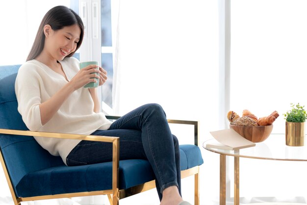 Relax and peaceful asian woman enjoy vacation weekend with hot tea and good book morning time near window home background