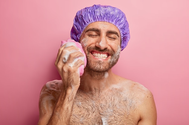 Relax, spa, hygiene, softness concept. Joyful smiling young man with broad smile, shows white perfect teeth, rubs cheek with sponge, has foam on body