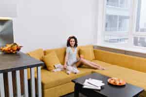 Бесплатное фото Отдыхайте в современной квартире счастливой молодой женщины, отдыхающей на оранжевом диване. журнал, чашка чая, домашние животные, радостное настроение, улыбка, настоящие эмоции
