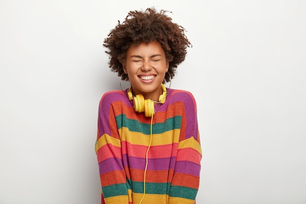 Relaxation and wellbeing concept. Overjoyed dark skinned woman enjoys perfect lazy weekend, keeps eyes shut from pleasure, dressed in oversized striped sweater, uses headphones