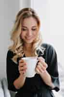 Free photo relaxed adorable smiling girl is smiling and holding morning cup of coffee indoor shot of amazing lady sitting at home and resting near the window