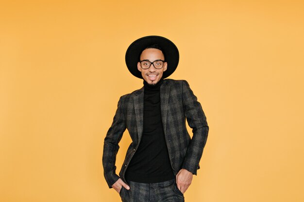 Расслабленный африканский парень в винтажном клетчатом костюме, улыбаясь на желтой стене. Возбужденный черный молодой человек в шляпе, весело во время фотосессии.