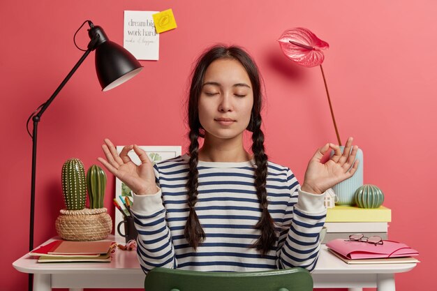 Relaxed Asian woman meditates at workplace, sits in zen pose against desktop with flowers, desk lamp, notepads, wears striped casual jumper, tries to relax after work, isolated over pink background