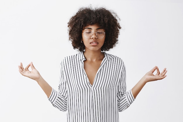 Relaxed attractive modern african american female businesswoman leading tiresome lifestyle gathering patience in yoga closing eyes, spreading hands in zen gesture