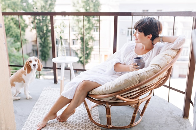 Relaxed barefooted girl in white dress sitting in chair on balcony and holding cup of tea