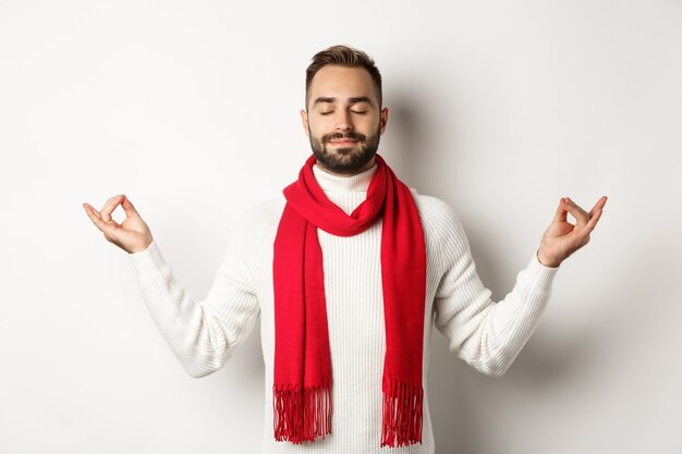 Relaxed bearded guy standing in peace, meditating with eyes closed, standing over white background in red scarf and sweater