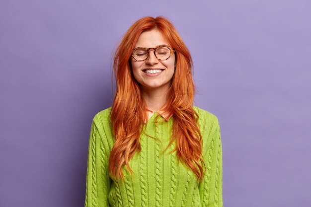 편안한 아름다운 빨간 머리 여자는 눈을 감고 미소는 녹색 점퍼와 안경을 착용하는 즐거운 말을 듣고 넓게 만족감을 느낍니다.