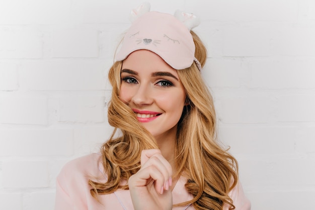 Relaxed blue-eyed woman in pink eyemask laughing on light wall. Photo of ecstatic girl with blonde wavy hairstyle wears sleepmask.