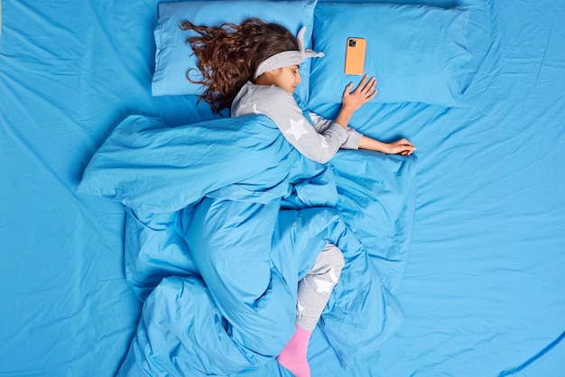 расслабленная брюнетка женщина, одетая в пижаму, покрытую мягким теплым пуховым одеялом, глубоко спит в спальне, позирует на кровати, мобильный телефон лежит рядом, есть некоторое время до будильника. Спокойный сон