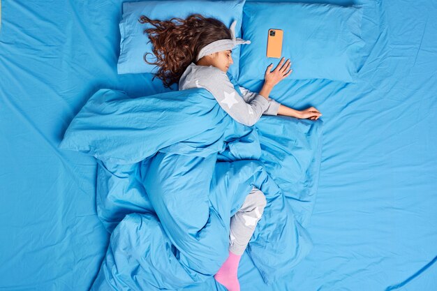 relaxed brunette woman dressed in pajama covered with soft warm duvet has deep sleep at bedroom poses on bed mobile phone lies near has some time before alarm clock. Peaceful nap