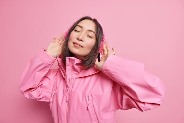 Relaxed carefree asian woman wears wireless headphones closes eyes enjoys favorite music cathes rhythm of song dressed in pink jacket poses indoor. monochrome shot. enjoying life during spare time