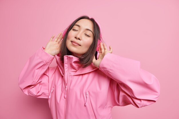 リラックスしたのんきなアジアの女性は、ワイヤレスヘッドホンを着用して目を閉じ、ピンクのジャケットのポーズに身を包んだ歌のリズムを室内で楽しんでいます。モノクロームショット。余暇の生活を楽しむ