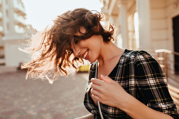 Relaxed caucasian girl waving her hair on the street. Smiling jocund woman in checkered shirt standing outdoor in autumn day.