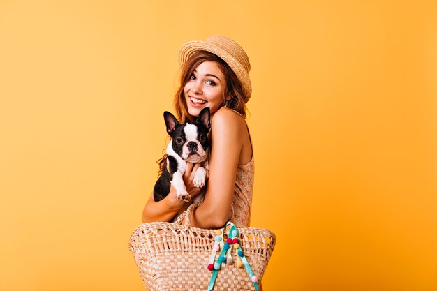 Relaxed caucasian lady embracing her cute dog. Excited red-haired girl in straw hat holding french bulldog.