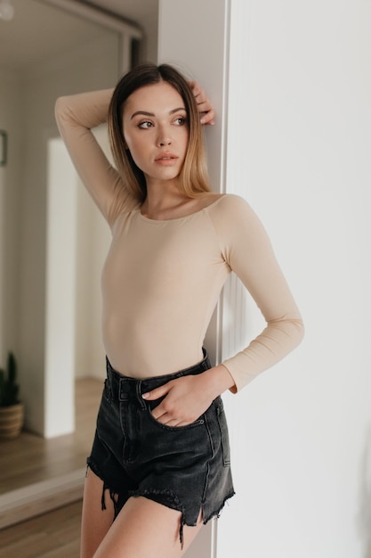 Free photo relaxed cheerful blond woman in stylish beige pullover and shorts smiles sincerely and looking aside happy pretty girl stands over white wall