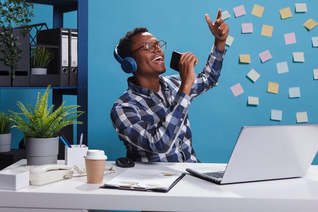 Relaxed company employee doing karaoke while sitting in office workspace. Cheerful smiling heartily young office worker wearing wireless headphones and listening to music.