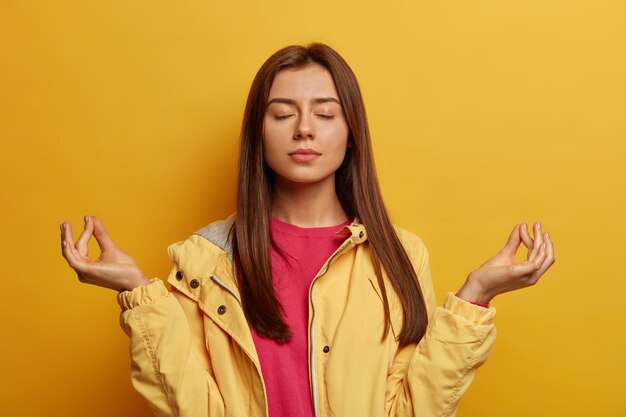 Relaxed dark haired young woman with healthy skin, makes zen mudra gesture, tries to calm down after hard day, breathes deeply and practices yoga, wears windbreaker, isolated on yellow wall