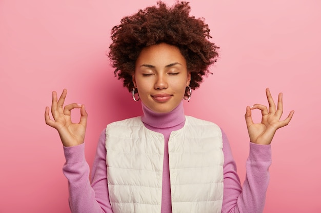 무료 사진 편안한 어두운 피부 소녀 인내심과 안도감, mudra zen 제스처를 보여주고, 흰색 조끼를 입은 후 요가 연습, 분홍색 배경에 눈을 감고 서 있습니다.