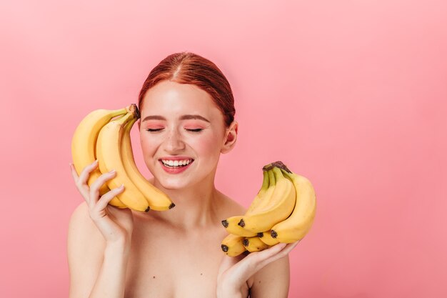 熟したバナナを持ってリラックスしたヨーロッパの女性。ピンクの背景に分離されたエキゾチックな果物でポーズをとる生姜の魅力的な女の子。