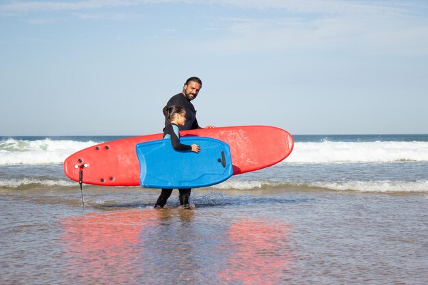 Relaxed father and daughter doing sports on beach. Mid adult man and dark-haired girl walking in water with surfboards. Family, leisure, active lifestyle concept