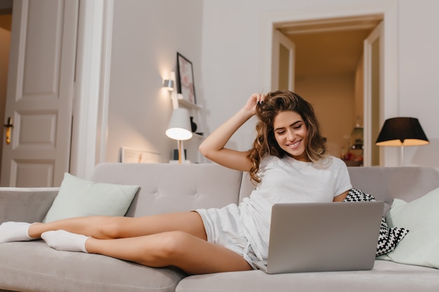 Relaxed female freelancer plays with her curly hair during work with laptop