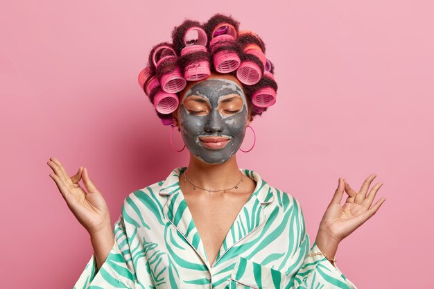 Relaxed female model meditates indoor makes zen gesture keeps eyes closed dressed in pajama applies beauty mask on face enjoys peacful domestic atmosphere isolated over pink wall. Facial care