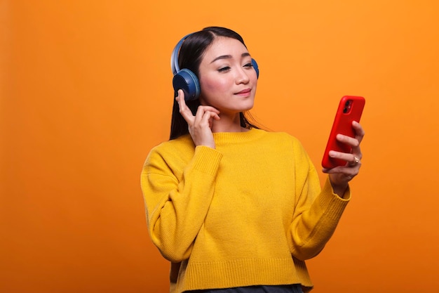 Relaxed friendly cute asian woman listening to music on audio streaming platform while using smartphone device. Calm person wearing headphones while enjoying leisure time by listening to song.