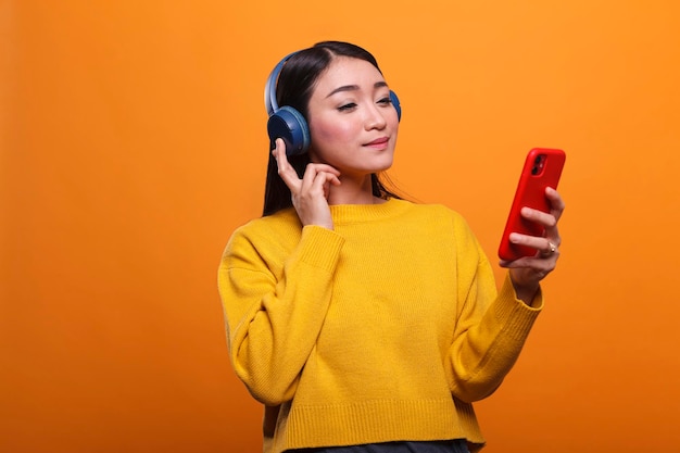 Расслабленная дружелюбная симпатичная азиатская женщина слушает музыку на платформе потокового аудио при использовании смартфона. Спокойный человек в наушниках, наслаждаясь досугом, слушая песню.