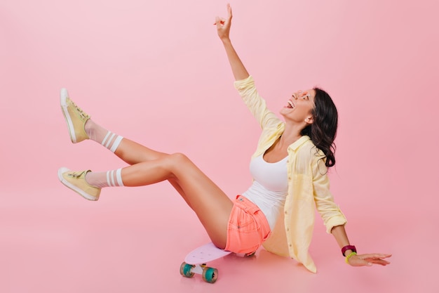 Relaxed girl in bright summer outfit sitting on skateboard with legs up and laughing. Beautiful young brunette lady in yellow shoes spending time with longboard.