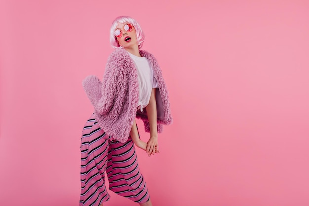 Relaxed girl in peruke funny posing on pink background Indoor shot of winsome female model wears striped pants and fur jacket