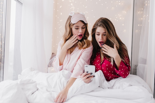 Relaxed girls expressing surprised emotions during photoshoot in bed. Indoor photo of amazed female friends sitting under white blanket with smartphone.