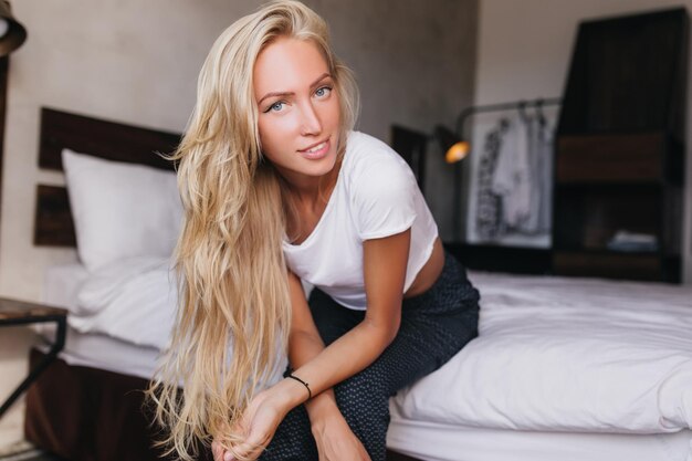 Relaxed gorgeous woman with long hairstyle smiling to camera Joyful blonde lady sitting on bed in room with cozy interior