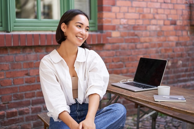 Free photo relaxed and happy asian woman sitting with laptop in outdoor cafe drinking coffee smiling and laughi