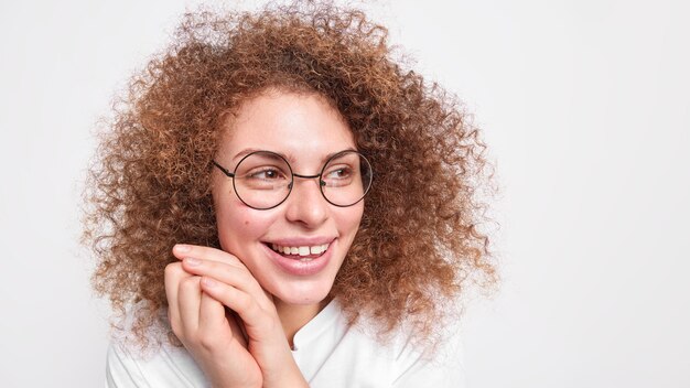 곱슬 덥수룩 한 머리카락을 가진 편안한 행복한 여자는 얼굴 미소 근처에 손을 유지하며 홍보를 위해 흰 벽 빈 복사본 공간에 대한 시력 교정 모델을위한 둥근 안경을 착용하고 좋은 하루를 즐깁니다.