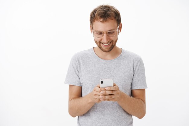 Relaxed and joyful handsome aduly male with beard in glasses and gray t-shirt looking down at smartphone screen with broad smile having fun messaging