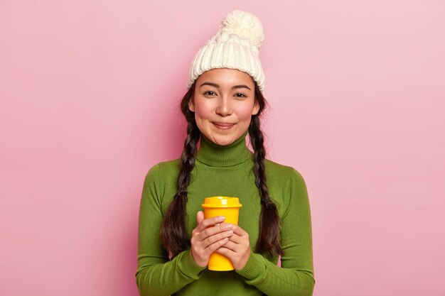 Relaxed lovely dark haired woman with two pigtails, holds takeout coffee, warms during winter day with hot beverage, wears white hat and green turtleneck