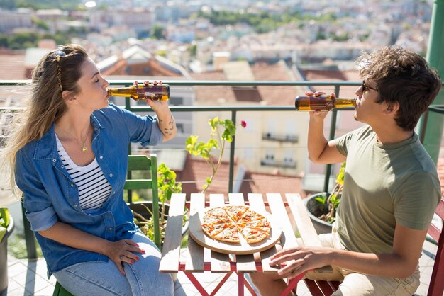 Relaxed male and female friends sitting at table at party. Young people in casual clothes sitting on terrace roof, talking, eating pizza and drinking beer. Communication, friendship concept