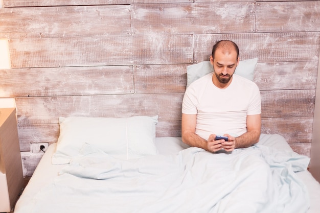 밤에 침대에 누워 휴대폰을 사용하는 편안한 남자.