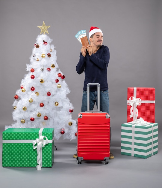 Free photo relaxed man with red suitcase showing his travel tickets on grey