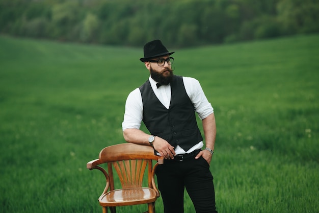 Uomo rilassato con gilet e cappello accanto a una sedia in legno