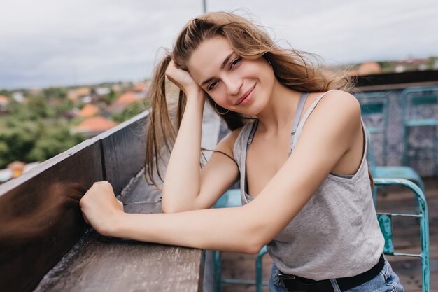 Relaxed pale woman sitting on roof in morning. Outdoor shot of enchanting long-haired girl posing with pleasure