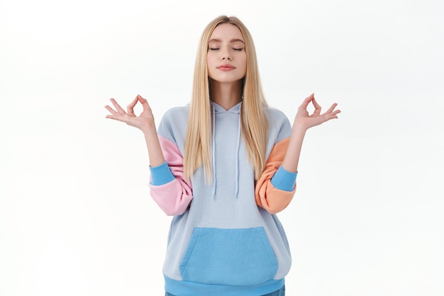 Free photo relaxed peaceful blonde girl in hoodie keep calm and patient close eyes and breathing during meditation reach zen or nirvana hold hands sideways standing in lotus pose white background