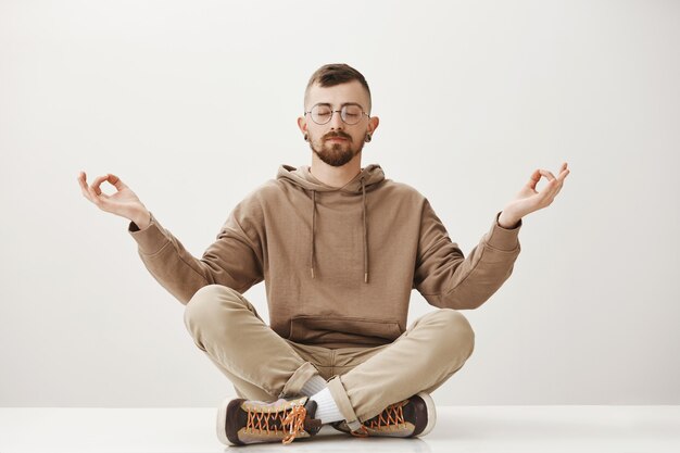 Расслабленный мирный хипстерский парень сидит на полу и медитирует, сохраняйте спокойствие