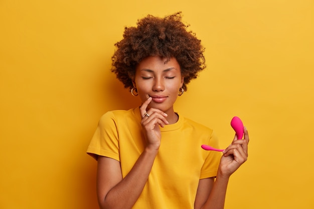 Relaxed pleased woman happy to buy smart vibrator, cant control vibrations speed via voice control, uses smartphone app, isolated on yellow wall. wireless remote control clitoris stimulator