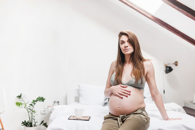 Relaxed pregnant woman with coffee mug sitting on bed