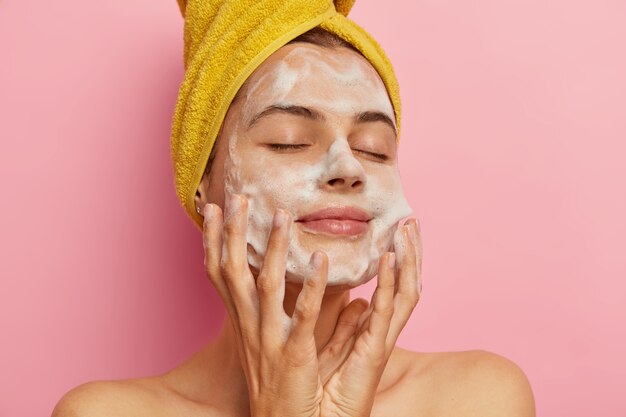 Relaxed pretty woman cares about her appearance, washes face with pleasant facial gel or soap, removes all pores, keeps eyes shut from pleasure, gets hygienic treatments