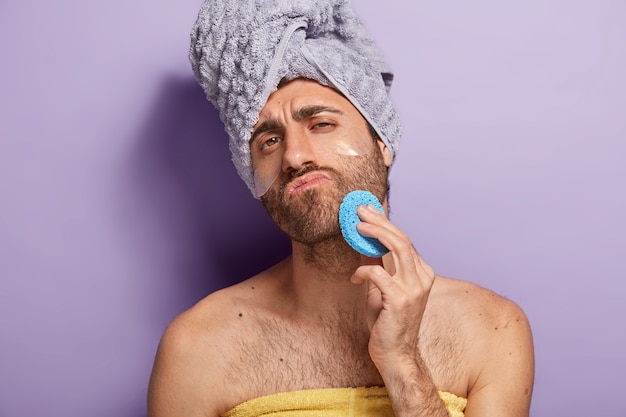 無精ひげでリラックスした真面目な男はシャワーの後に顔の皮膚を拭き、化粧用スポンジを保持し、柔らかいタオルで包んだ