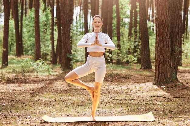 Relaxed sporty woman standing in open air with closed eyes keeping palms together, standing on one leg, wearing sportswear, enjoying training in beautiful forest.