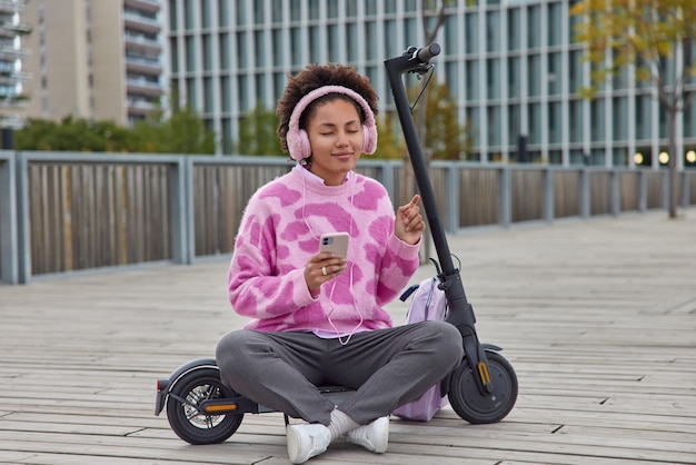 Relaxed teenage girl sits crossed legs near electric scooter enjoys favorite music in headphones rests after ride dressed casually poses in urban place prefers eco transport. Millennial lifestyle